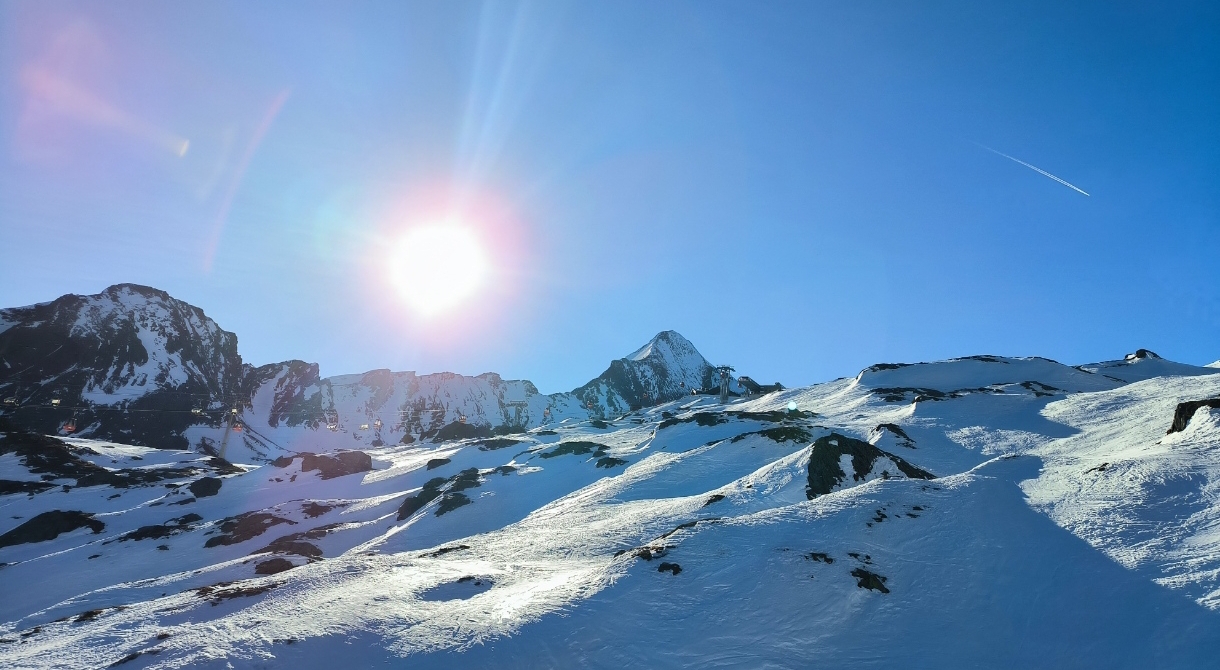 Das Bild zeigt eine schneebedeckte Berglandschaft unter klaren blauem Himmel. Die Sonne ist nahe der Bildmitte deutlich zu erkennen. Die Berggipfel sind etwas zerklüftet.